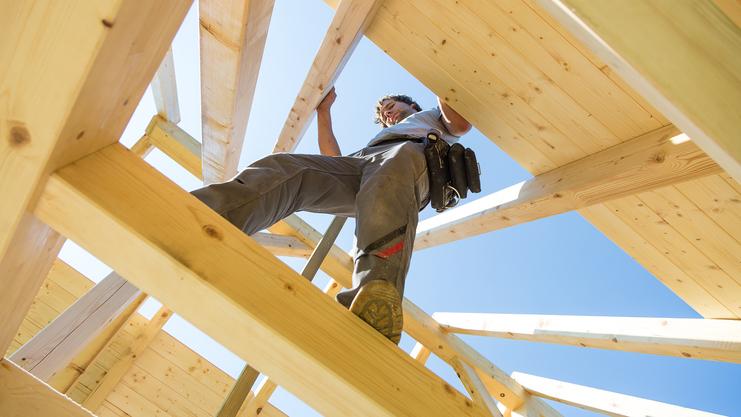 Blick von unten auf einen Handwerker, der an einer Dachkonstruktion aus Holz arbeitet, bei klarem blauen Himmel.